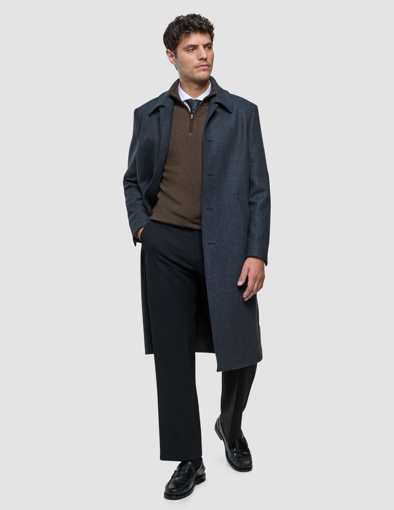 Ein Mann, der einen dunkelgrauen Maxi-Wollmantel über einem braunen Pullover mit Viertelreißverschluss, einem weißen Hemd mit Kragen, einer Krawatte, einer schwarzen Hose und Loafern trägt.